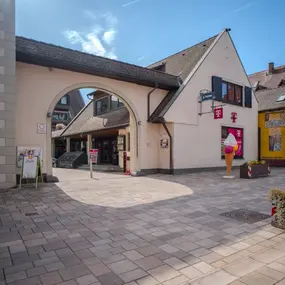 Fußgängerzone mit Telekom-Shop, Eisdiele und Blumenbeeten bei sonnigem Wetter.